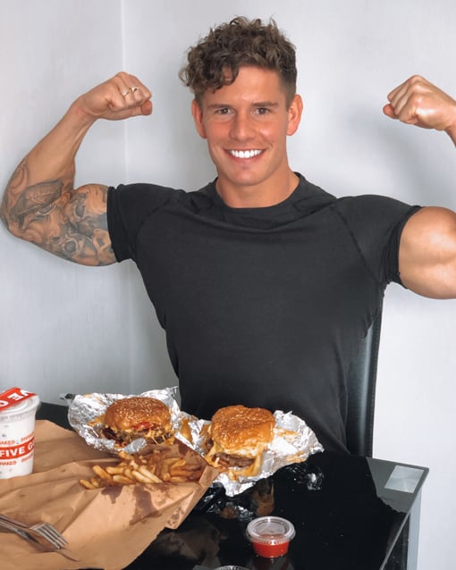 Luke grinning and flexing both arms behind two burgers and a tray of fries