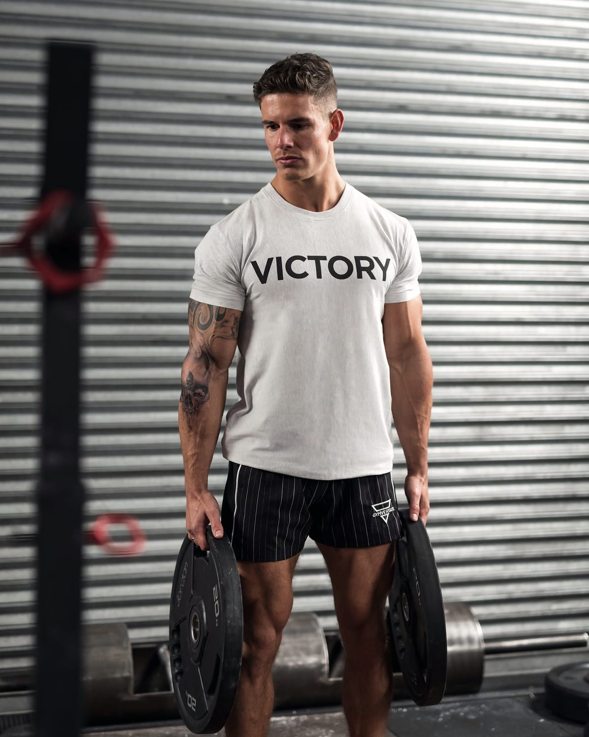 Luke Hayes in a white VICTORY T-shirt holding two weight plates in front of a roller shutter