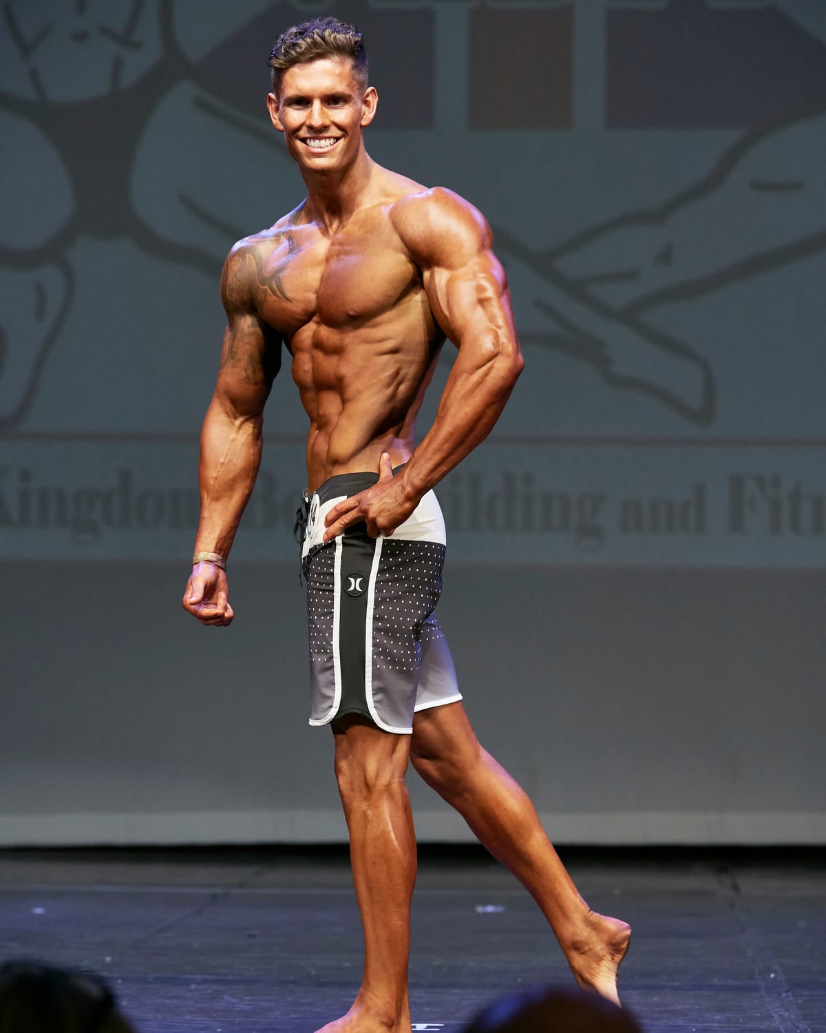 Luke Hayes on stage at a UKBFF physique competition, smiling mid-pose