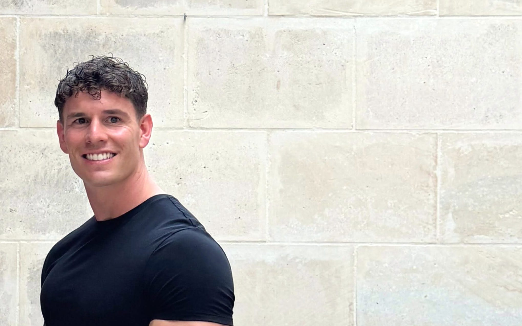 Luke Hayes smiling in a black T-shirt against a pale stone wall